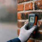Diagnostic d’humidité sur façade en briques à Bruxelles : main gantée avec humidimètre (19,6 %) sur mur présentant zones sombres et efflorescences.